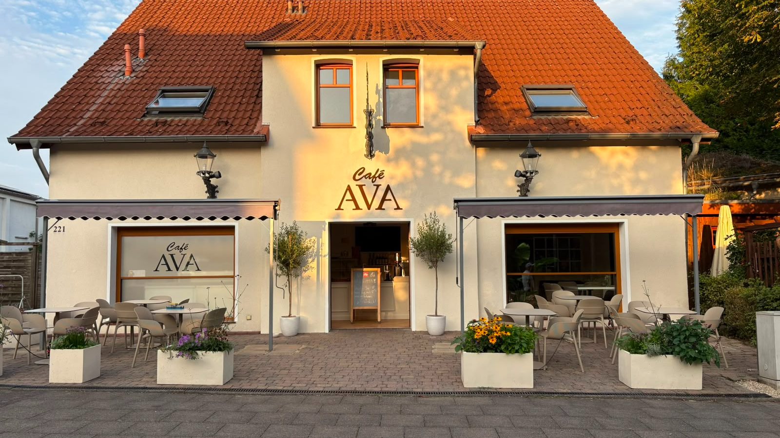 Café Ava von außen: helles Haus mit rotem Ziegeldach und Terrasse mit Tischen im Abendlicht